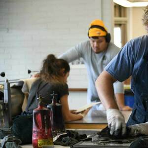 A smiling young man with blonde hair wears a paint-splattered blue apron and white gloves while cleaning a printing plate. Two other students, one in a yellow beanie, work nearby in an art studio. Red bottles are on the table.